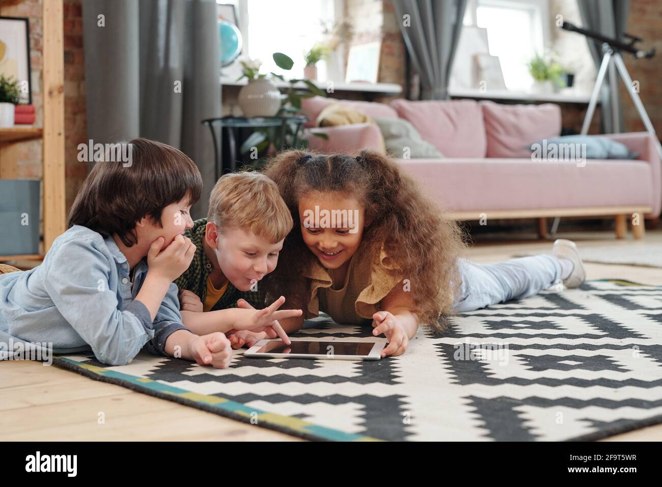 Gruppo di bambini felici che giacciono sul pavimento e giocano in un tablet digitale insieme nella stanza di casa Foto Stock