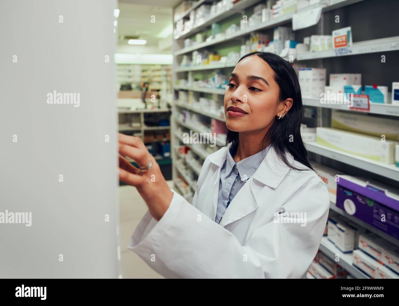 Giovane donna farmacista che indossa un camice da laboratorio alla ricerca di medicina in scaffale in farmacia Foto Stock