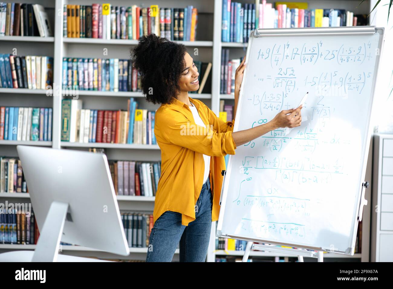 Formazione a distanza, lezione online. Una giovane insegnante afroamericana intelligente conduce lezioni online per gli studenti tramite videoconferenza utilizza il computer, si trova in biblioteca, mostra informazioni sulla lavagna Foto Stock