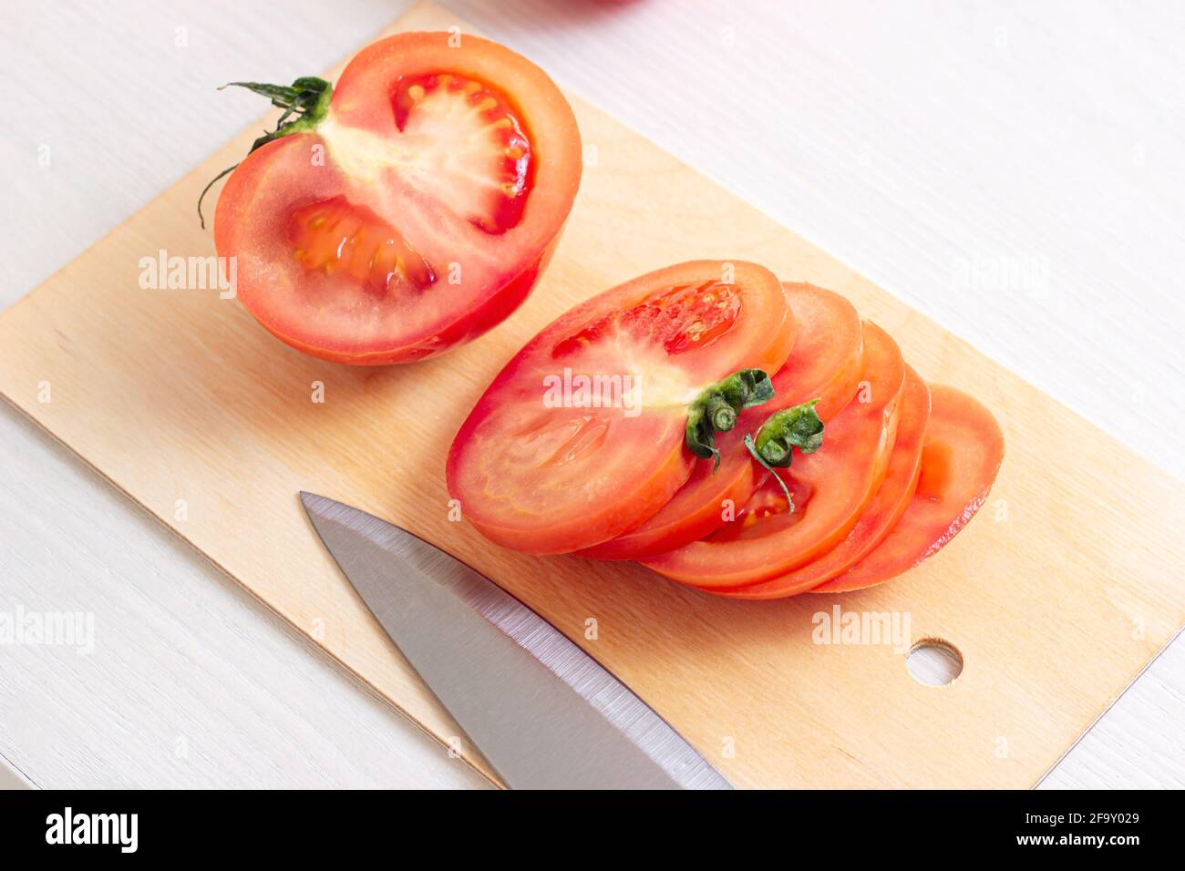 Pomodori rossi crudi freschi e brillanti tagliati con coltello su tagliere in legno in cucina su sfondo chiaro. Foto Stock