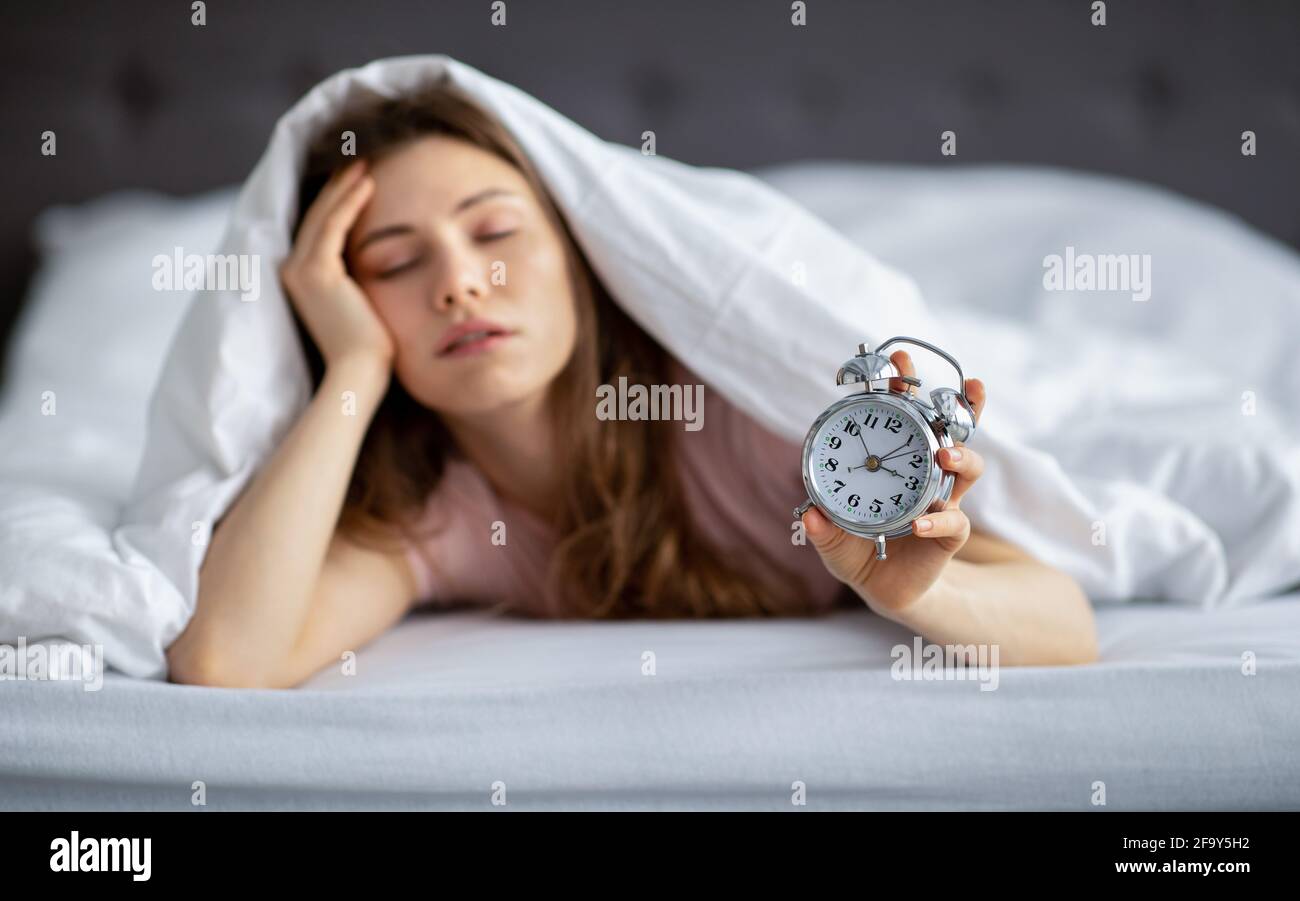 Sonnolenta giovane donna sdraiata sotto coperta a letto con sveglia, avendo problemi a svegliarsi al mattino, al chiuso Foto Stock