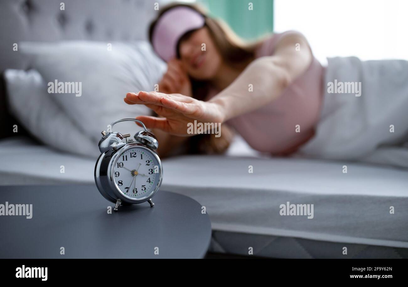 Giovane donna in maschera di sonno che spegne la sveglia al mattino, fuoco selettivo Foto Stock