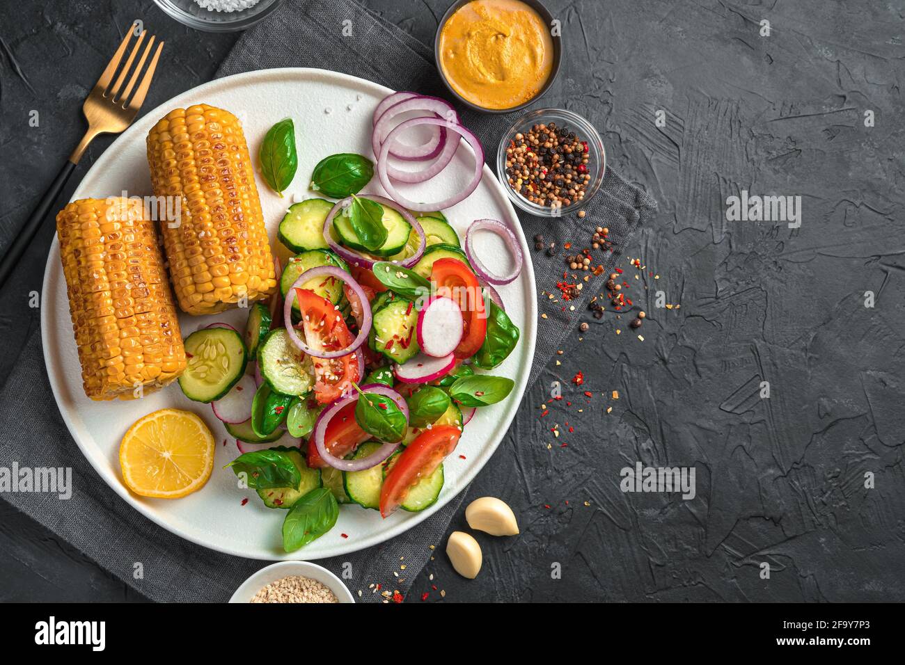 Insalata di verdure con pomodori, cetrioli e ravanelli e teste di mais in un piatto bianco piatto su fondo nero con spazio da copiare. Il concetto di Foto Stock