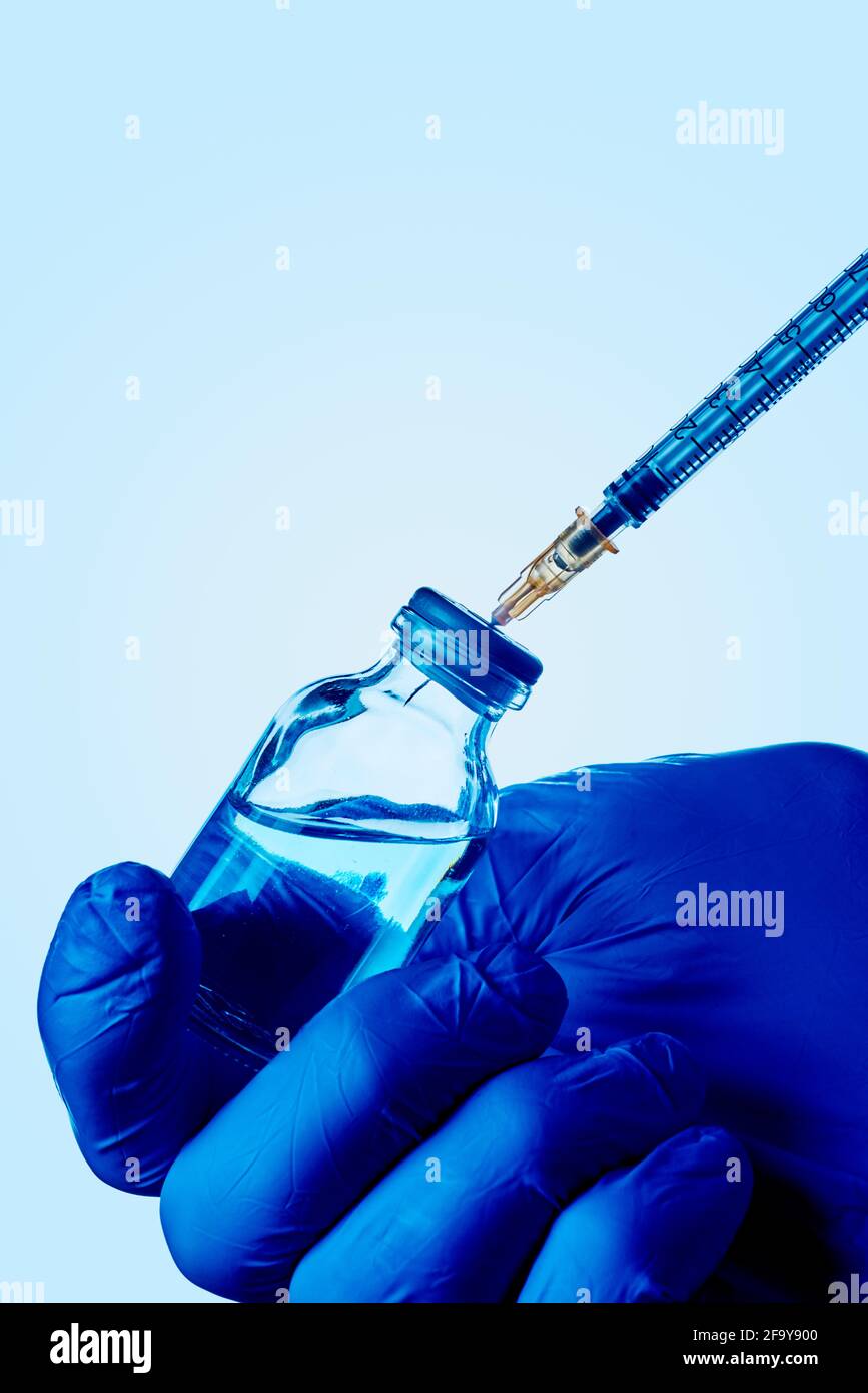 un operatore sanitario o di laboratorio, indossando guanti chirurgici blu, riempie una siringa di un flaconcino con un liquido blu Foto Stock