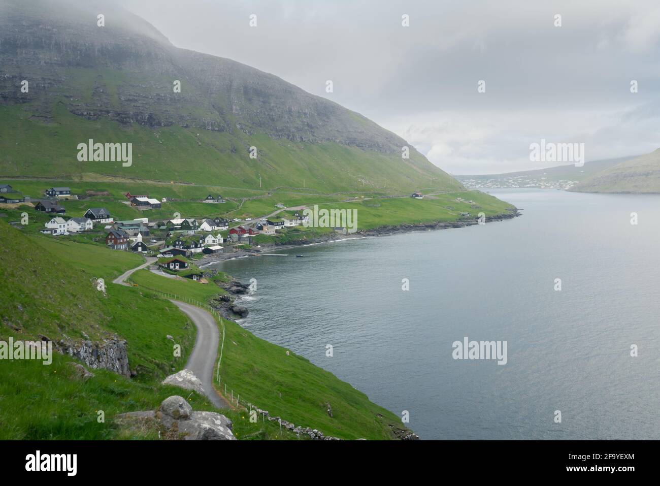 Una strada che conduce al villaggio faroese di Bour in una giornata fredda e nuvolosa. Erba verde su ripide scogliere e fiordo oceanico. Paesaggio faroese selvaggio. Foto Stock