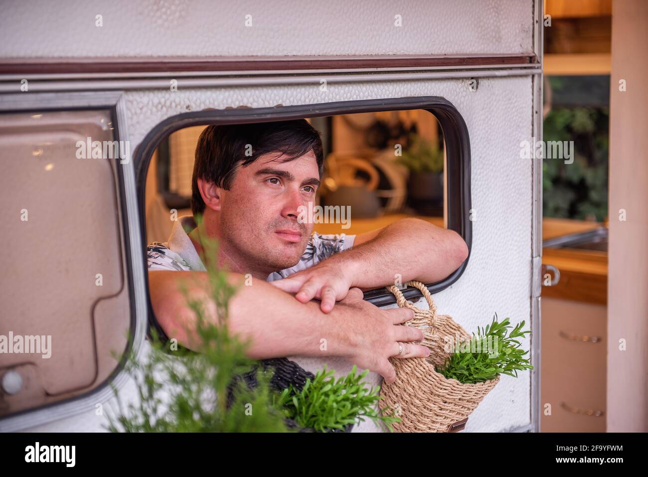 Ritratto di un giovane guarda fuori la finestra di una casa di camion rimorchio tra piante verdi. La brunetta riposa con la sua famiglia fuori città sulla t Foto Stock