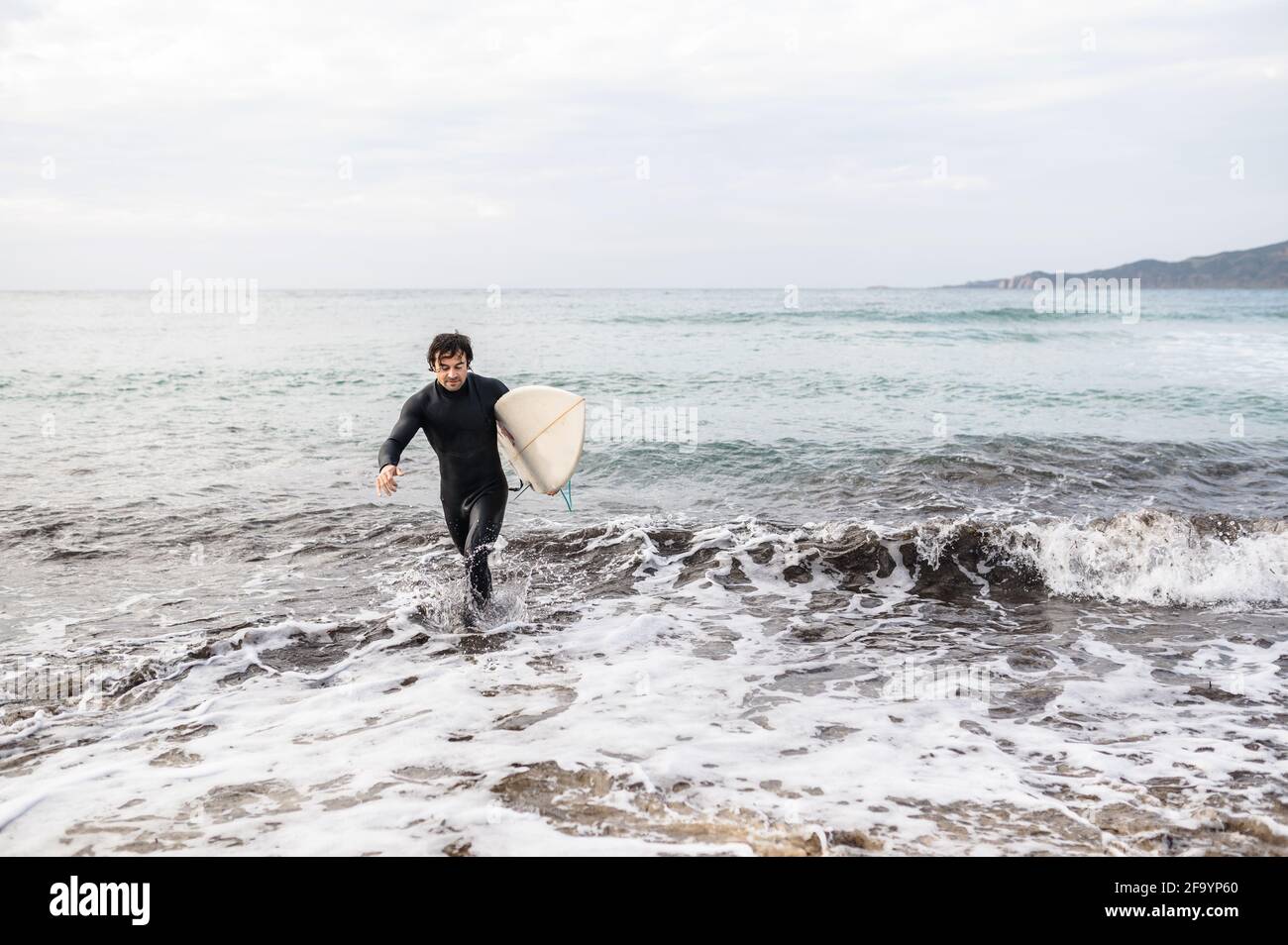Ritratto dell'uomo surfista che porta con sé una tavola da surf e indossa un costume da immersione che si allontana dall'acqua dopo il surf. Foto Stock