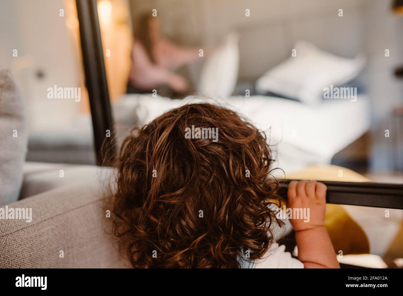 Adorabile bimbo con lunghi capelli ricci che guardano attraverso una finestra di una stanza alla madre Foto Stock