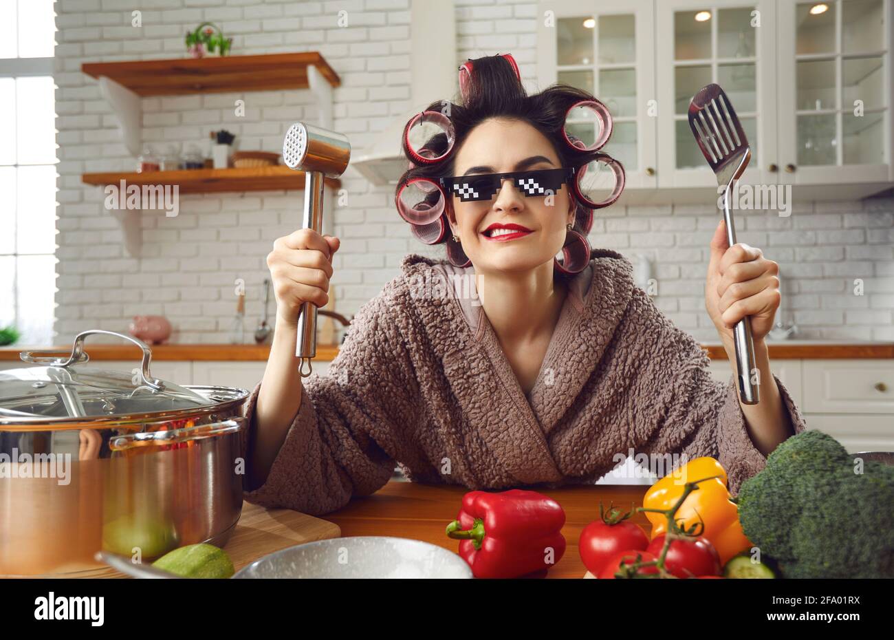 Casalinga divertente in arricciacapelli e bicchieri di vita thug che tengono la spatola e martello di carne Foto Stock