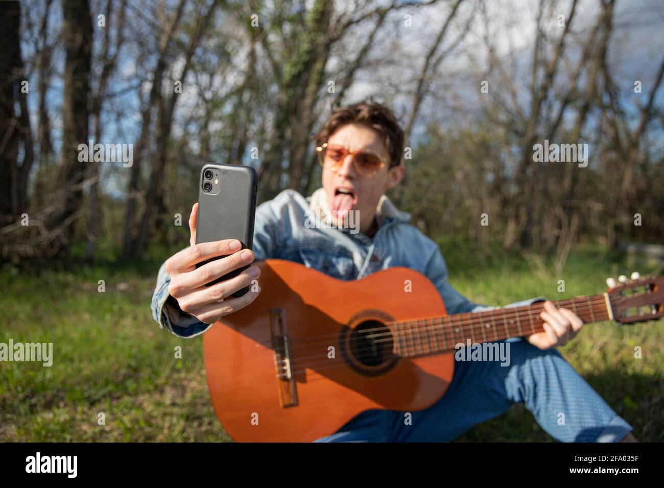 Primo piano della mano di un giovane musicista che prende un selfie mentre suona la chitarra, fa una faccia divertente con la sua lingua fuori Foto Stock