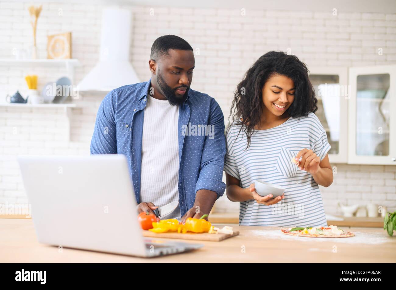 Eccitata giovane coppia afroamericana seguendo i passi da guardare le lezioni di cucina online, imparare a fare la pizza, guardare video blog corso dal portatile in cucina, il posizionamento di verdure Foto Stock