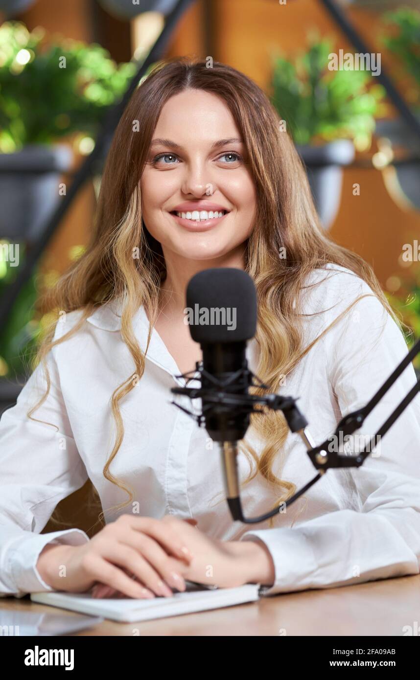 Primo piano ritratto di sorridente donna carina con capelli lunghi in camicia bianca che comunica con i seguaci online con il moderno microfono nero. Concetto di processo che parla blogger e abbonati online. Foto Stock