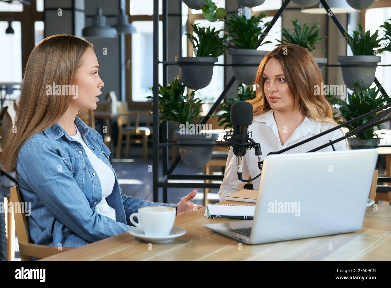 Vista laterale di due giovani blogger donne che comunicano su diversi argomenti con il moderno laptop e microfono. Concetto di sedersi in un bar e parlare in trasmissione da laptop per i seguaci. Foto Stock