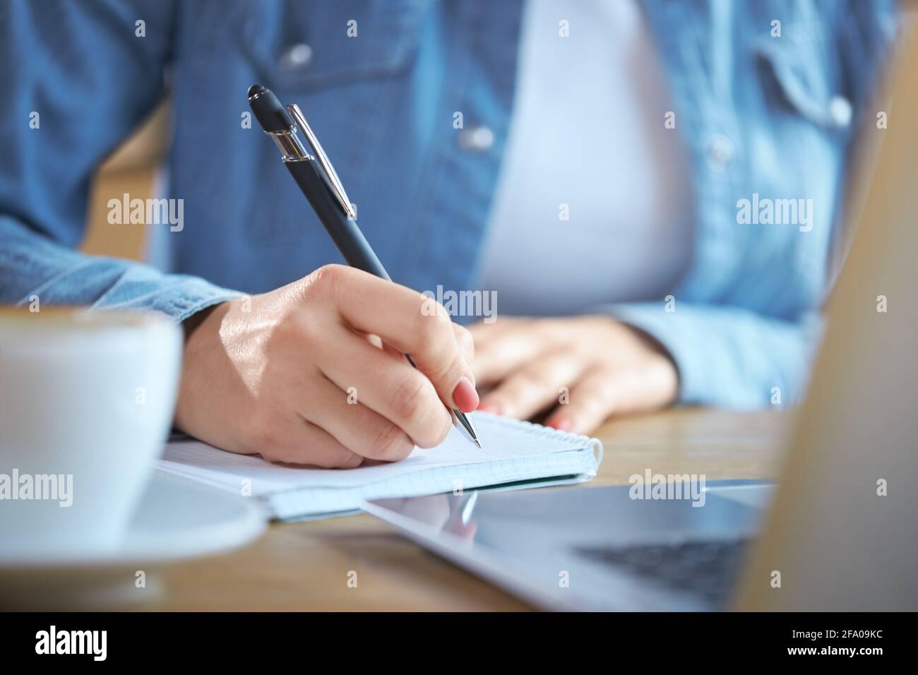 Primo piano di donna che tiene la penna e scrive alcune informazioni interessanti nel notebook. Concetto di processo preparazione per la comunicazione online con gustoso caffè e computer portatile moderno. Foto Stock