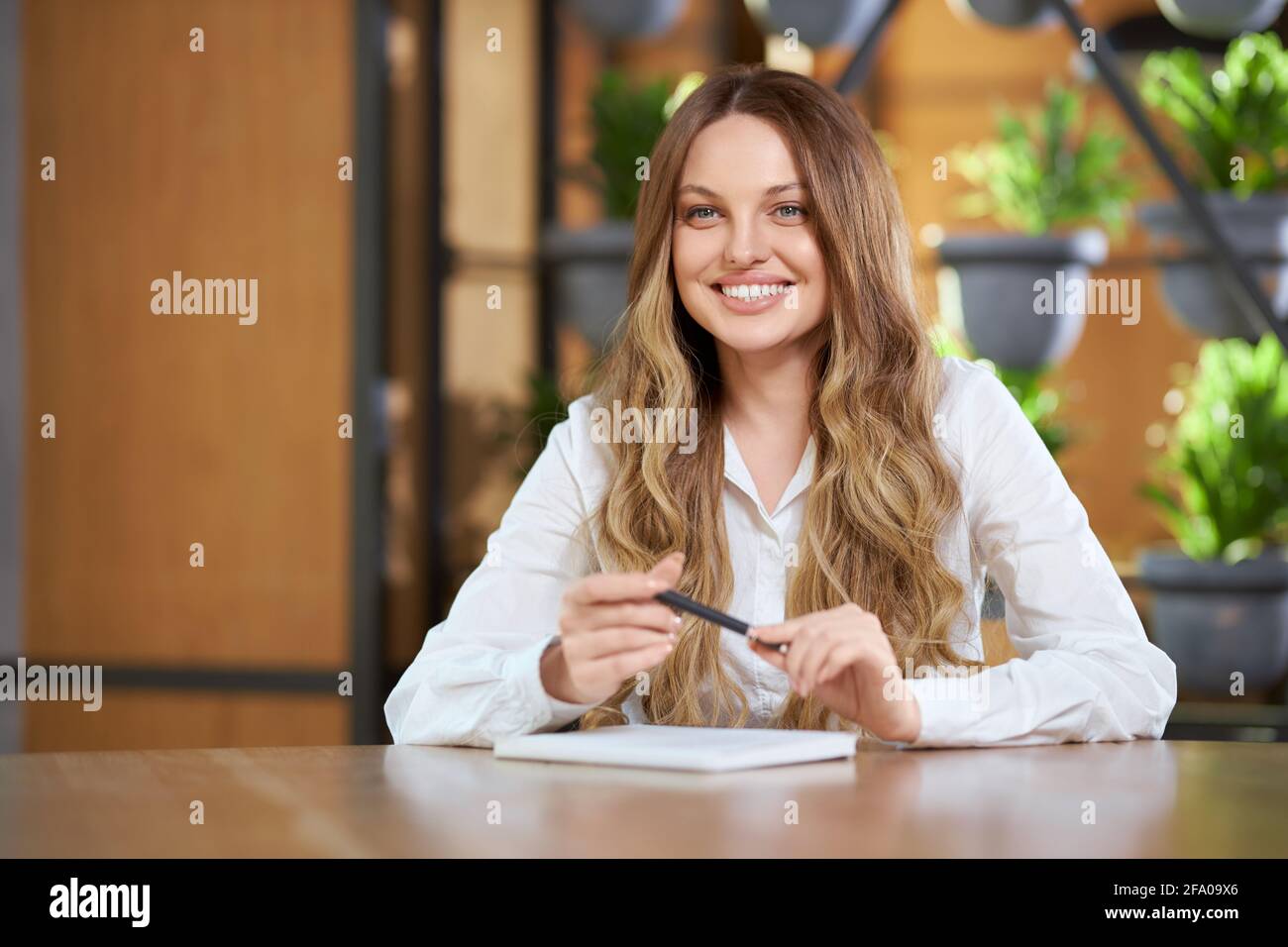 Primo piano ritratto di giovane donna sorridente con capelli lunghi in camicia bianca seduta al tavolo e scrivendo informazioni diverse. Concetto di colloquio di processo o di comunicazione in caffetteria. Foto Stock