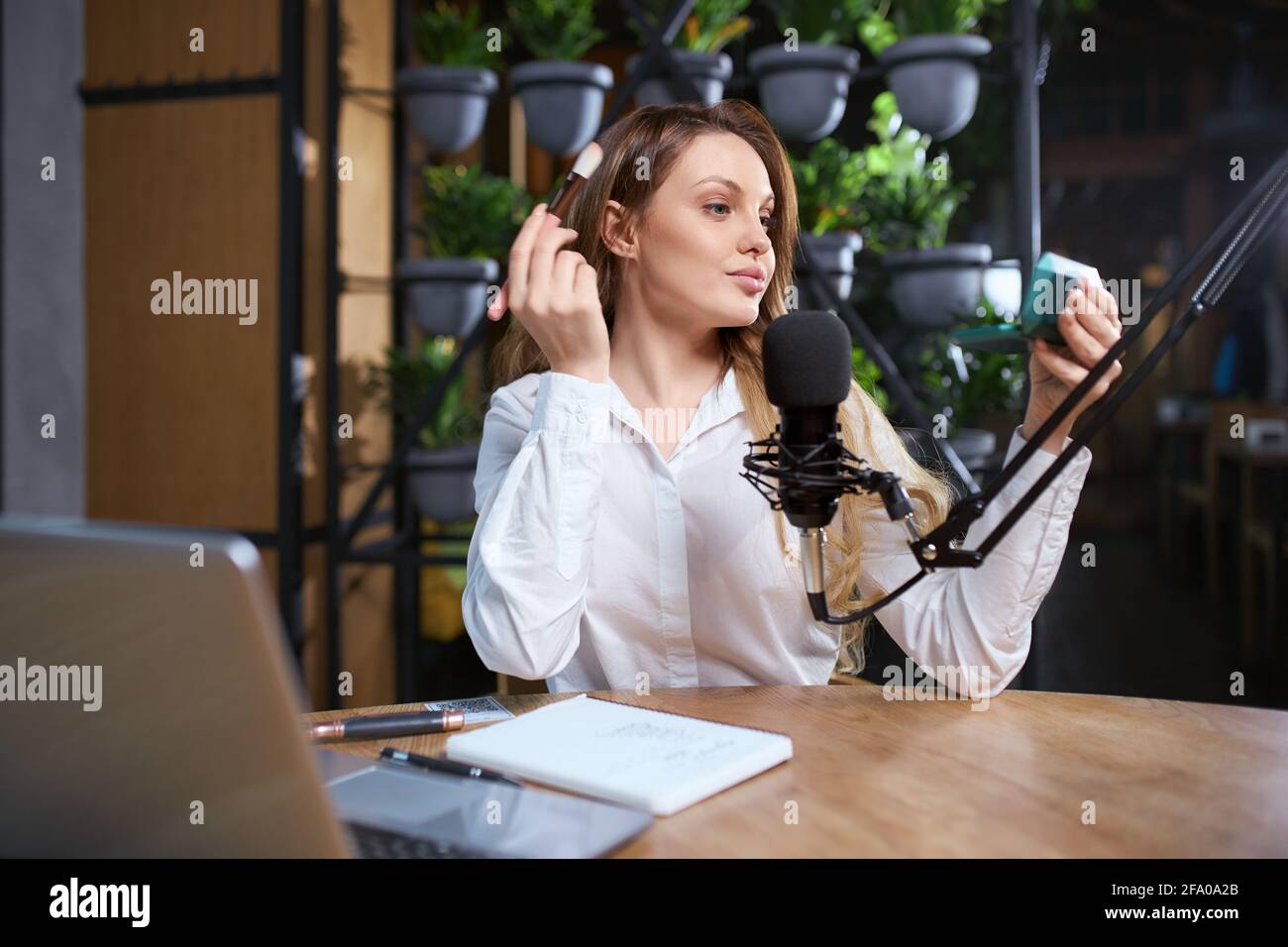 Vista frontale ritratto di attraente giovane donna che si prepara per l'intervista e facendo trucco con cosmetic. Concetto di sedersi al tavolo vicino al moderno microfono nero e al computer portatile. Foto Stock