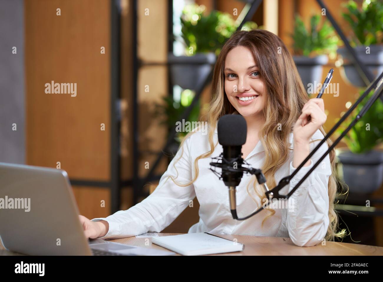 Primo piano di sorridente blogger donna in camicia bianca seduta al tavolo e comunicando con i seguaci online dal computer portatile. Concetto di processo live broadcast con microfono speciale moderno. Foto Stock