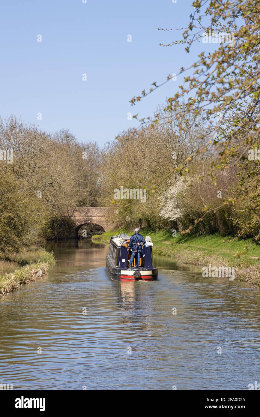 Crick, Northamptonshire, Regno Unito - 19 aprile 2021: Un uomo e una donna su una narrowboat che naviga verso un ponte sul Canal Grande Union in primavera. Foto Stock