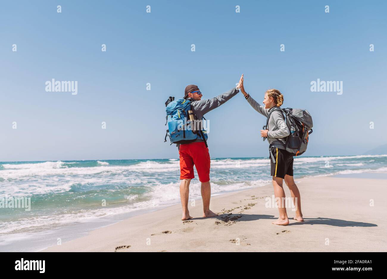 Padre e figlio adolescente con zaini che fanno High FIVE sulla spiaggia sabbiosa di mare durante la passeggiata di trekking Lycian Way. Famoso percorso turco Likya Yolu. ACTIV Foto Stock