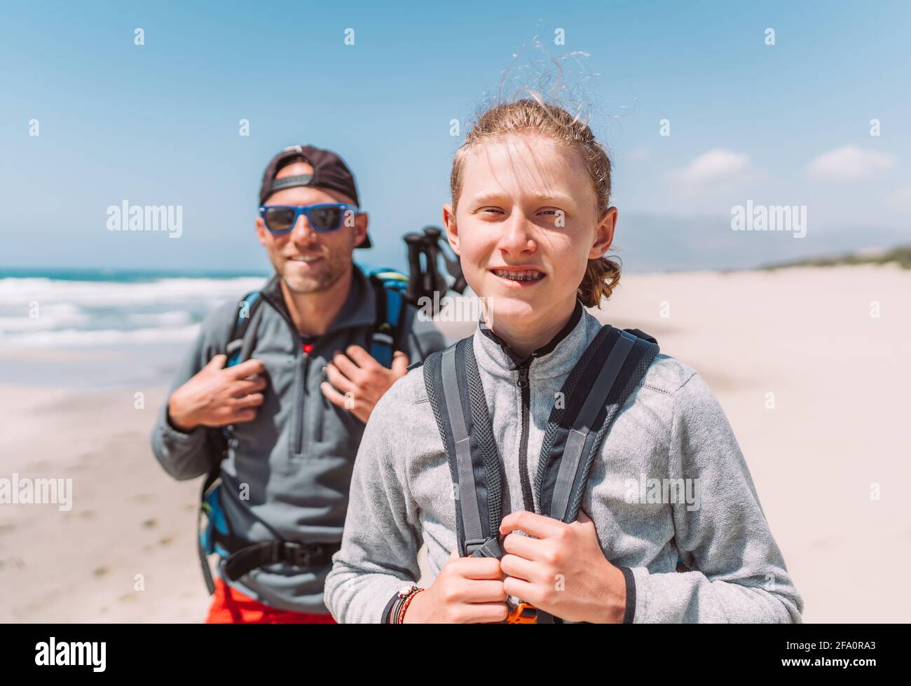 Padre con figlio adolescente con zaini a piedi dalla spiaggia di sabbia. Sorridono e guardano la fotocamera. Attiva felice famiglia persone vacatio Foto Stock