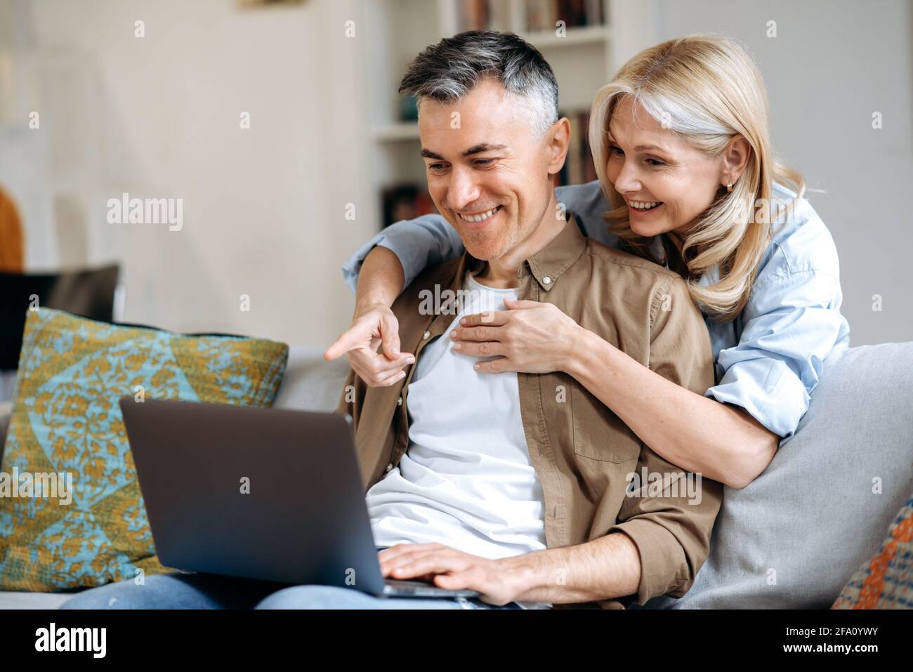 Gioiosa coppia caucasica matura trascorrere il tempo a casa, seduto sul divano in un abbigliamento casual elegante, utilizzando il computer portatile per conferenze online con amici o famiglia, navigare in internet, gesturing e sorridente amichevole Foto Stock