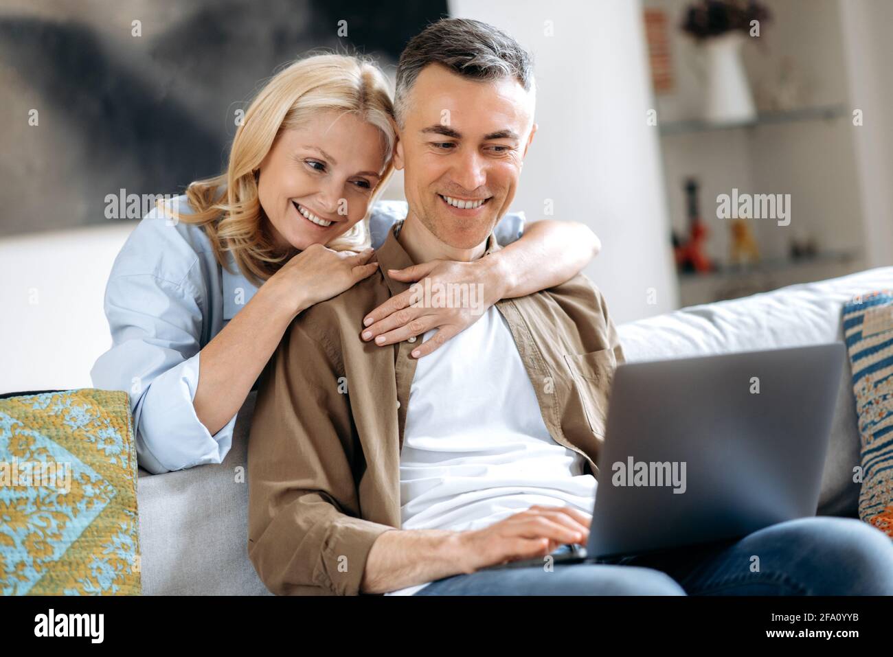 Gioiosa coppia caucasica adora a casa sul divano in abiti casual eleganti, utilizzando il laptop, navigando sui social media, chattando con amici o familiari, facendo acquisti online, abbracciando e sorridendo Foto Stock