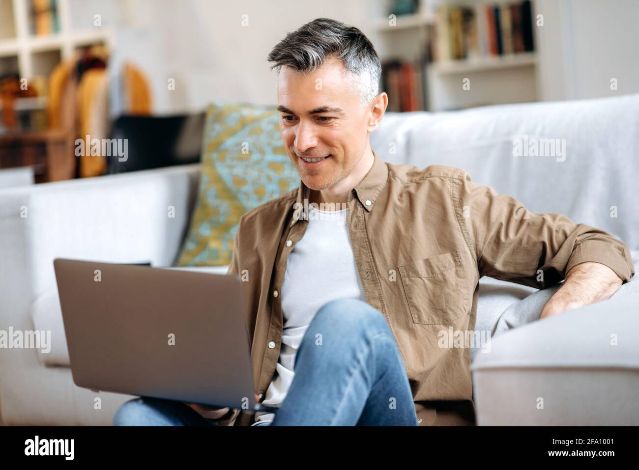 Bell'uomo caucasico dai capelli grigi senior seduto a casa in abiti casual eleganti, usa un laptop, SMS con amici o familiari, navigare in rete sociali, cercare informazioni, sorrisi Foto Stock