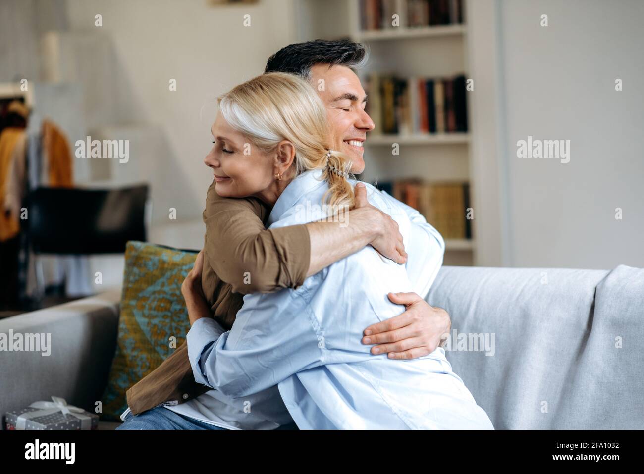 Felice amorevole famiglia caucasica senior, marito felice e moglie vestita in abiti casual elegante, seduto a casa sul divano in soggiorno, abbracciando, occhi chiusi, passare il tempo insieme, sorridendo Foto Stock