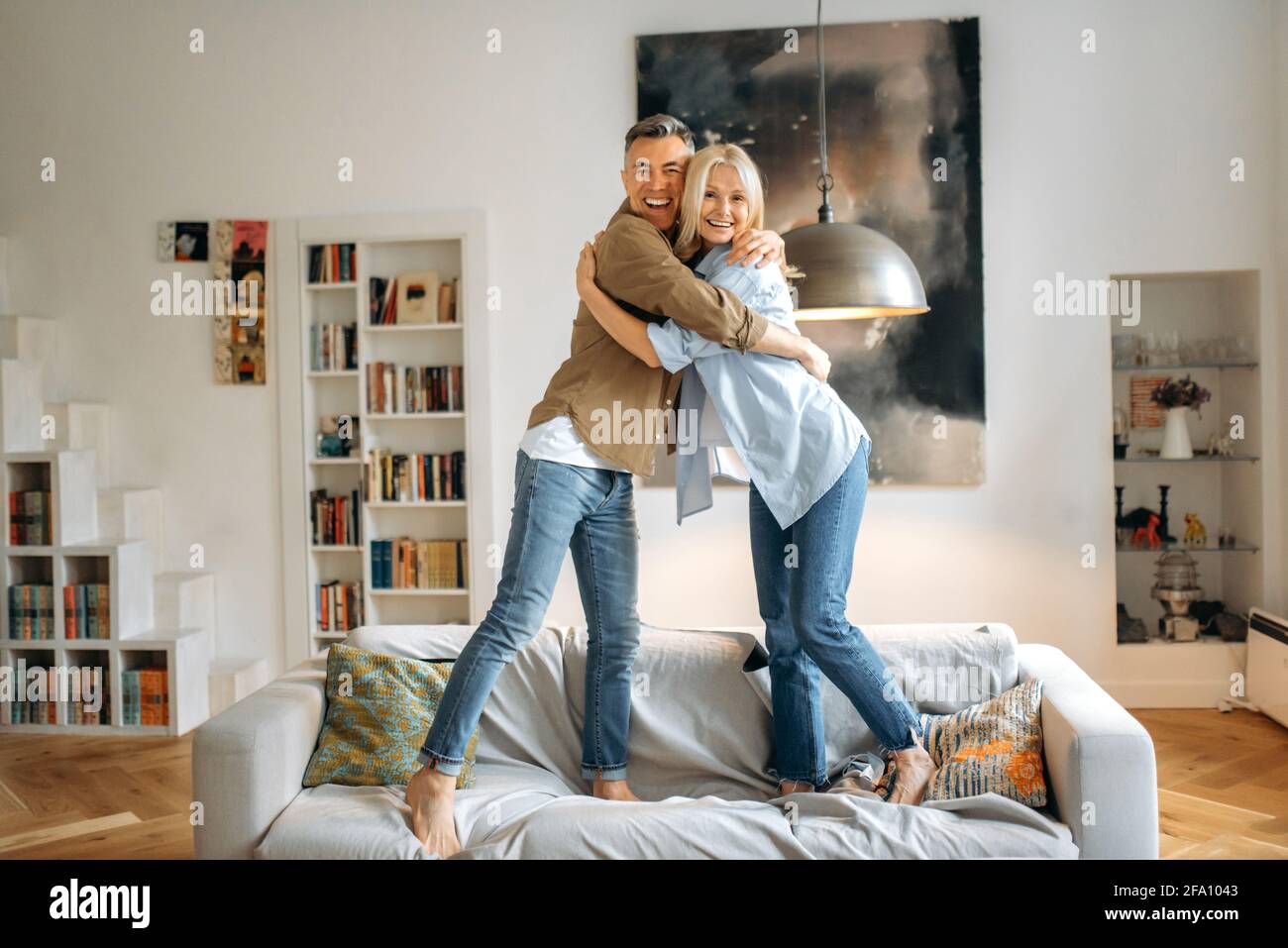 Felice carino gioioso maturo caucasico sposato coppia vestito in abiti casual elegante, marito e moglie sono in piedi sul divano a casa, abbracciando, sorridendo, guardando la fotocamera, divertendosi insieme Foto Stock