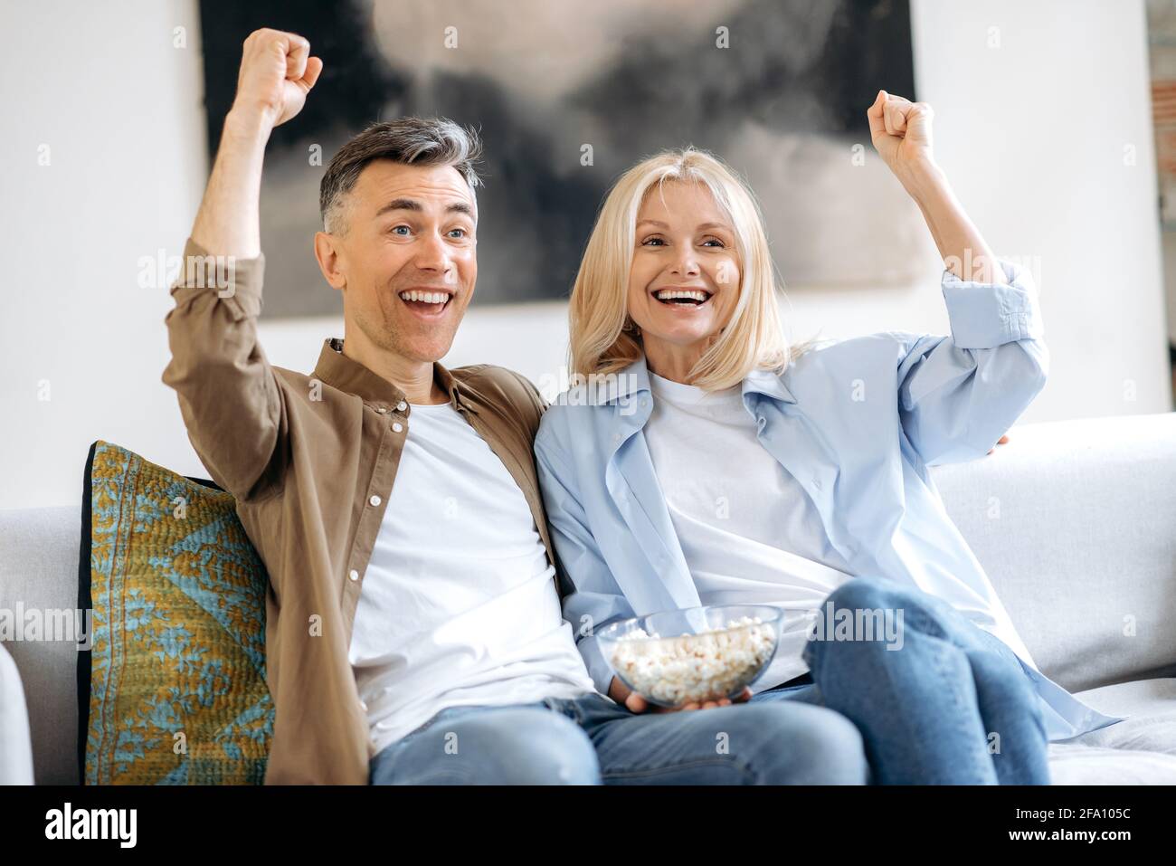 Gioiosa coppia sposata caucasica di mezza età, marito e moglie, seduta a casa sul divano in abiti casual eleganti, guardando la TV show, film o calcio, mangiare popcorn, gesturing, gioia di vincere Foto Stock