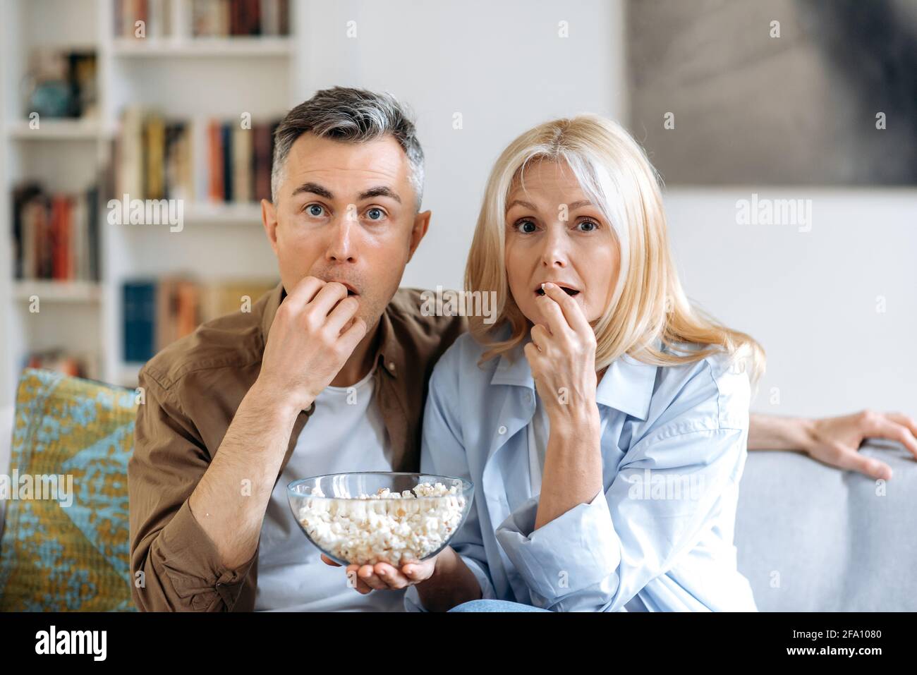 La coppia di famiglia caucasica di mezza età è rimasta scioccata e si siede a casa su un divano in abiti casual, guardando uno spettacolo televisivo o un film emozionante, mangiando popcorn, trascorrendo un fine settimana insieme a casa Foto Stock