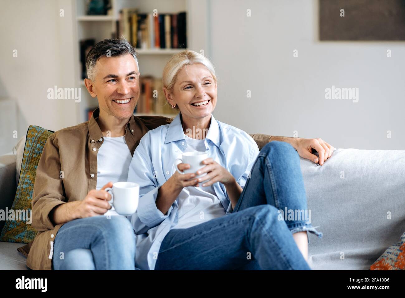 Felice coppia sposata caucasica matura in abiti eleganti casual, seduta su un comodo divano nel soggiorno, tenendo tazze di tè, chiacchierando, trascorrendo il tempo a casa, guardando via, sorridendo Foto Stock