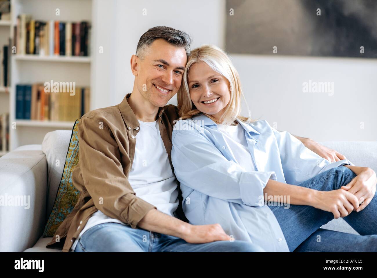 Ritratto di una piacevole coppia di sposi caucasici felici e maturi che si rilassano sul divano nel soggiorno di casa, trascorrendo il tempo insieme, vestiti in abiti casual eleganti, sorridendo alla macchina fotografica, felici insieme Foto Stock