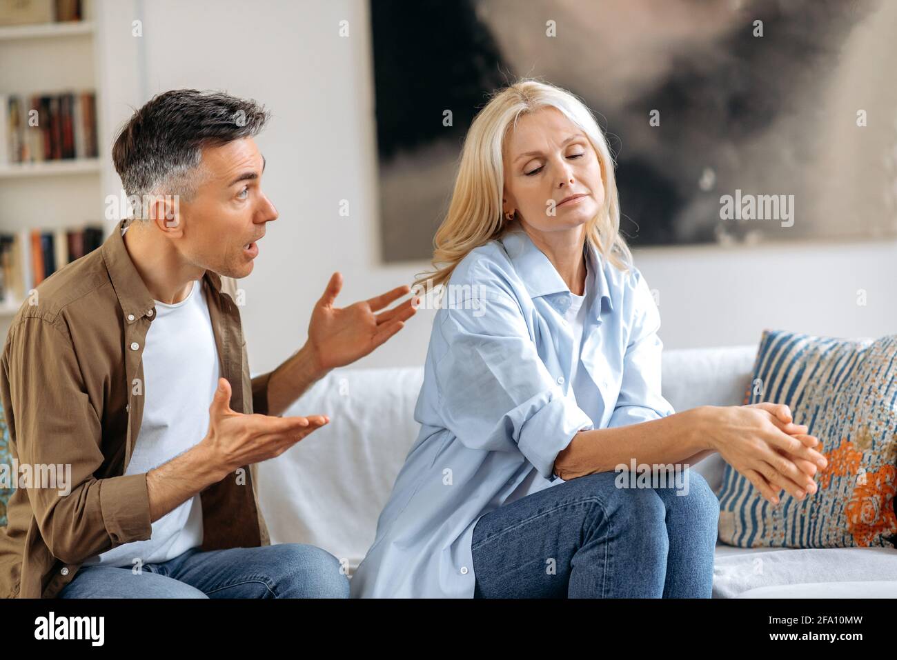 Incomprensioni, lite familiare. Sconvolto, indignato marito dai capelli grigi sta parlando emotivamente e guardando sua moglie, gesturando con le mani, moglie è sconvolto a causa di un dialogo, sperimentando stress Foto Stock