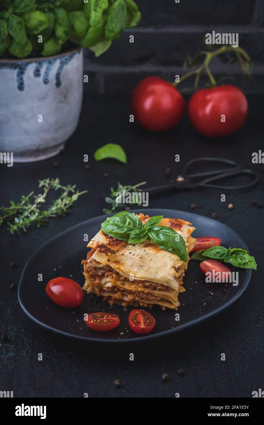 Pezzo di lasagna di soia vegetariana fatta in casa su un piatto nero, pomodori, erbe e basilico su sfondo scuro, verticale Foto Stock