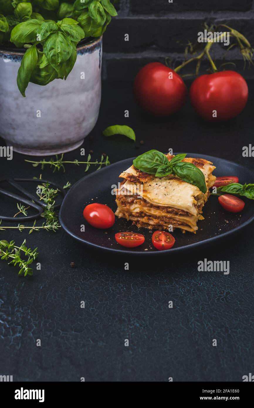 Pezzo di lasagna di soia vegetariana fatta in casa su un piatto nero, pomodori, erbe e basilico su sfondo scuro, verticale con spazio copia Foto Stock