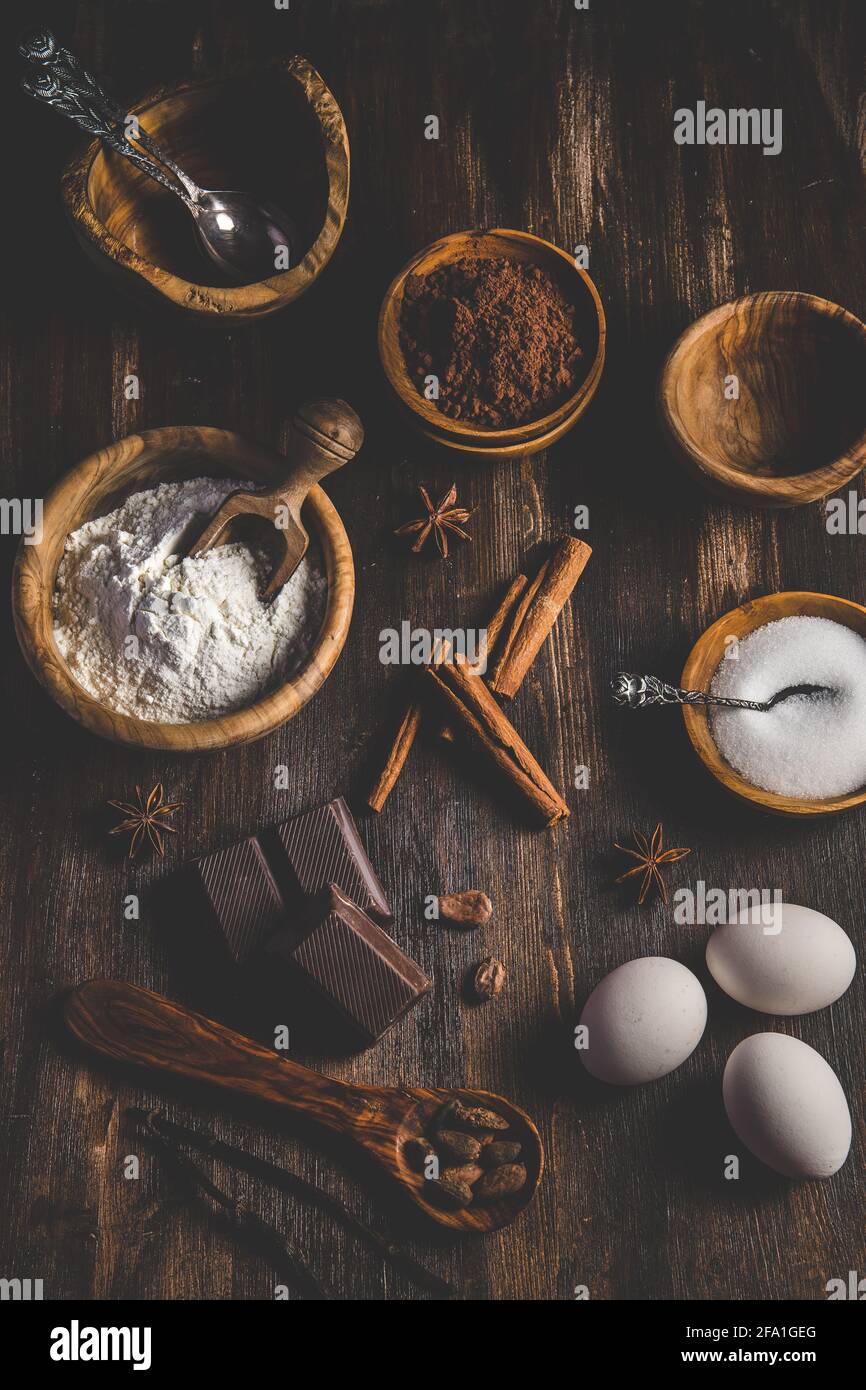 Vari ingredienti da forno come farina, uova, zucchero e cacao in ciotole di legno su un tavolo di legno scuro, vista dall'alto verticale Foto Stock
