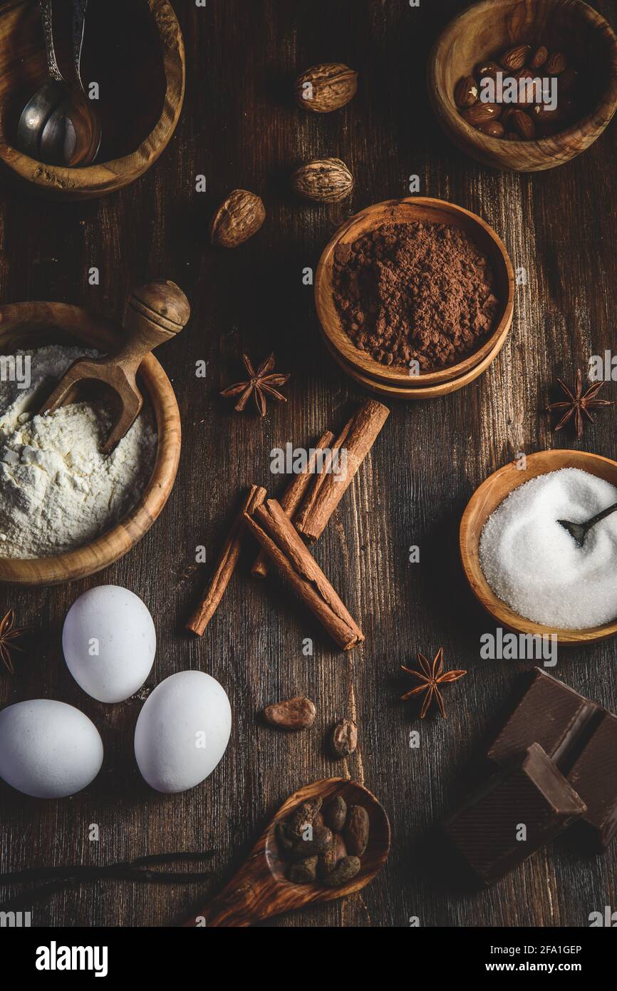Vista dall'alto verticale di vari ingredienti da forno come farina, uova, zucchero e cacao in ciotole di legno su un tavolo di legno scuro Foto Stock