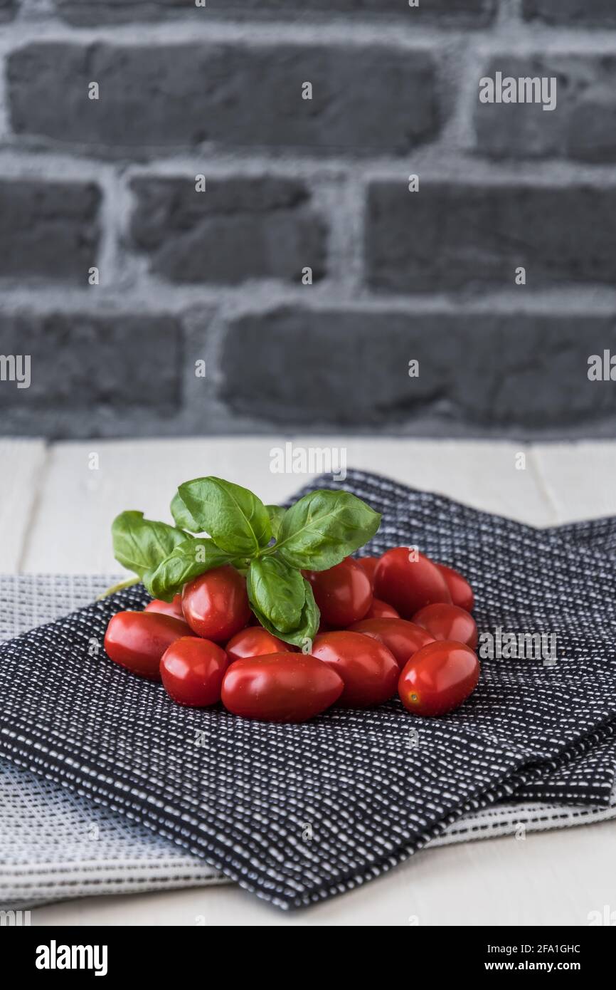 Pomodori ciliegini maturi con ramificazione di basilico su tovaglioli da tè bianchi e neri su sfondo grigio di pietra. Foto verticale. Foto Stock