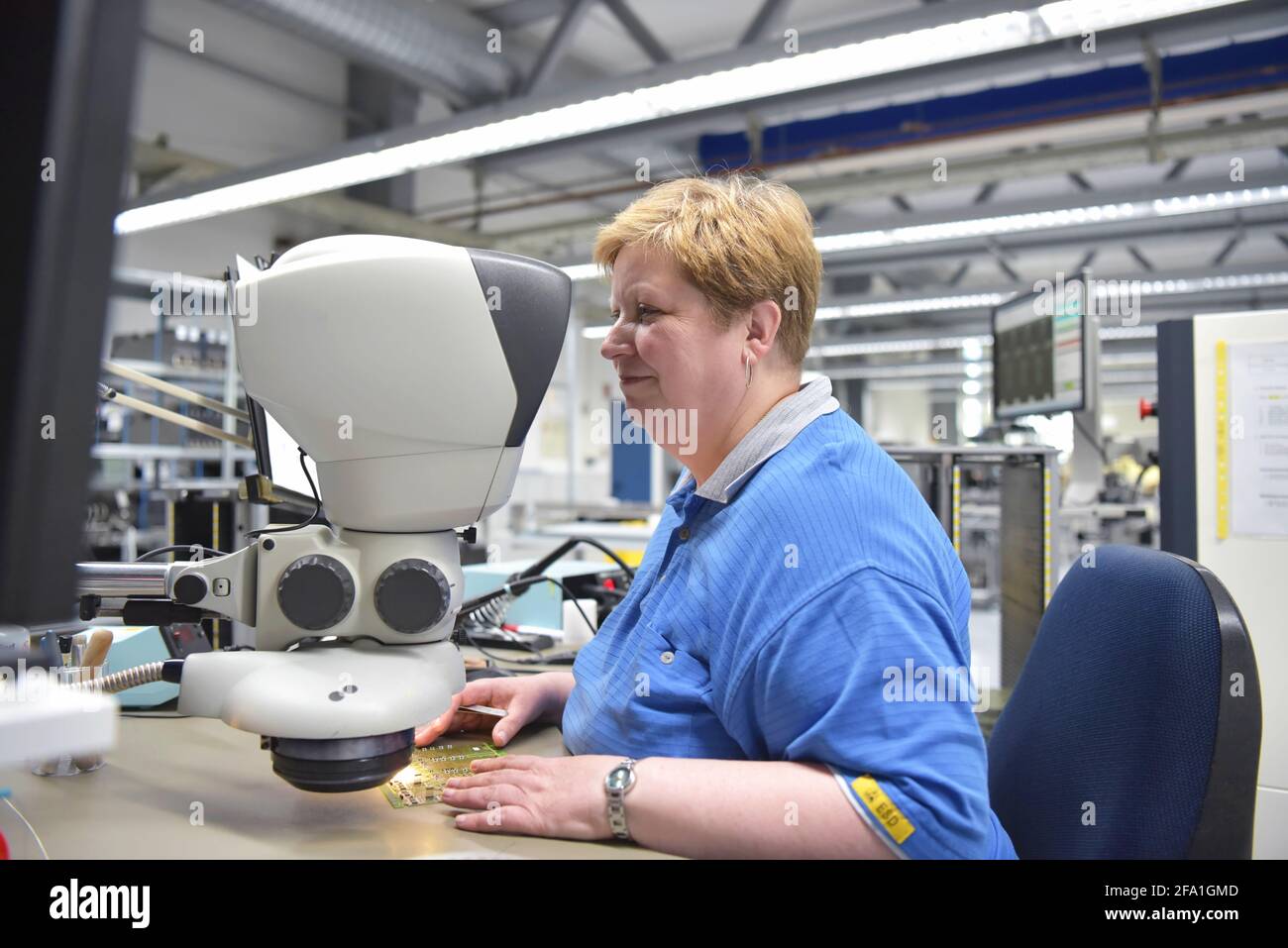 La donna in una fabbrica per la produzione di componenti elettronici controlla la qualità di una scheda assemblata con l'aiuto di un microscopio o ingrandimento di gl Foto Stock