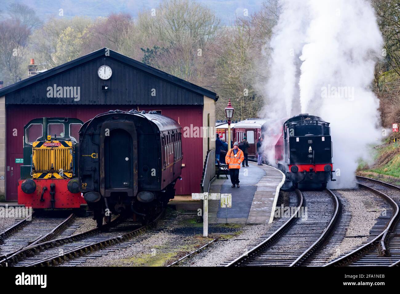 Treno a vapore storico o luogo che soffia nuvole di fumo (persone su piattaforma, operaio ferroviario, capannone motore) - Oxenhope Station sidings, Yorkshire, Inghilterra Regno Unito. Foto Stock