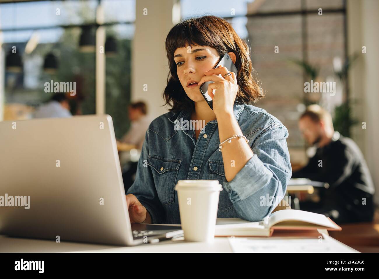 Freelance che lavora su un computer portatile e parla su un telefono cellulare in uno spazio di co-working. Donna che fa affari in uno spazio di co-working. Foto Stock