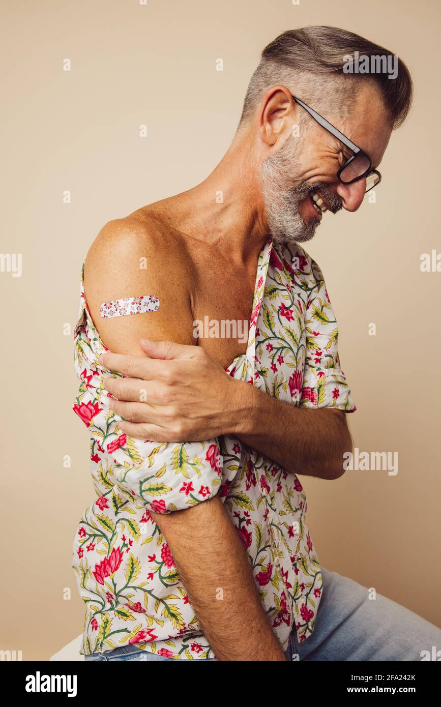 Uomo felice seduto con bendaggio sul braccio dopo aver ottenuto la vaccinazione. Uomo maturo che mostra il braccio dopo aver ricevuto il vaccino covid-19. Foto Stock