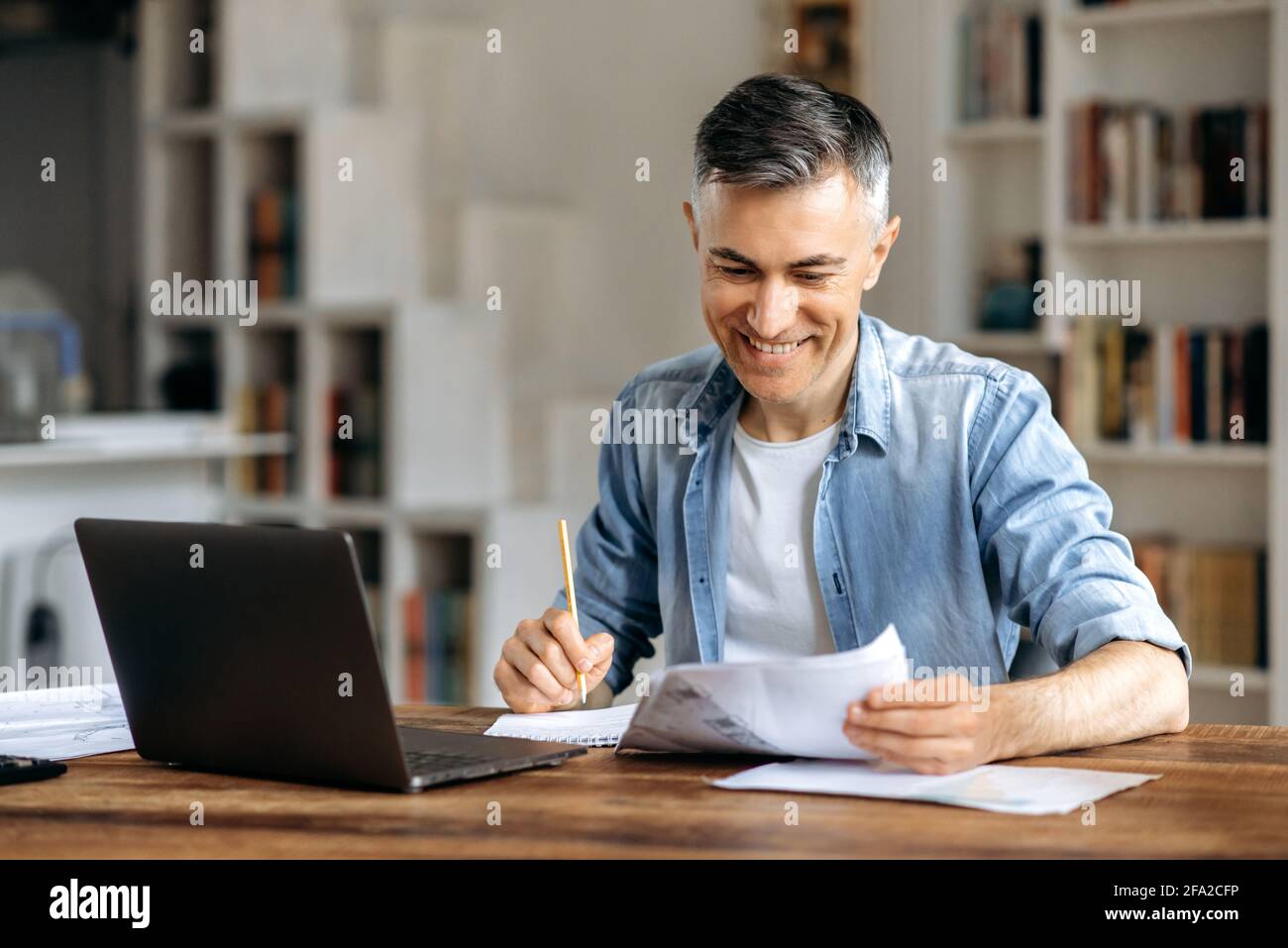 Soddisfatto riuscito sicuro maturo grigio-capelli caucasici uomo d'affari, manager o freelancer, in abbigliamento casual elegante, lavorare in ufficio utilizzando il laptop, controllare i documenti di lavoro, prendere appunti, sorridere Foto Stock