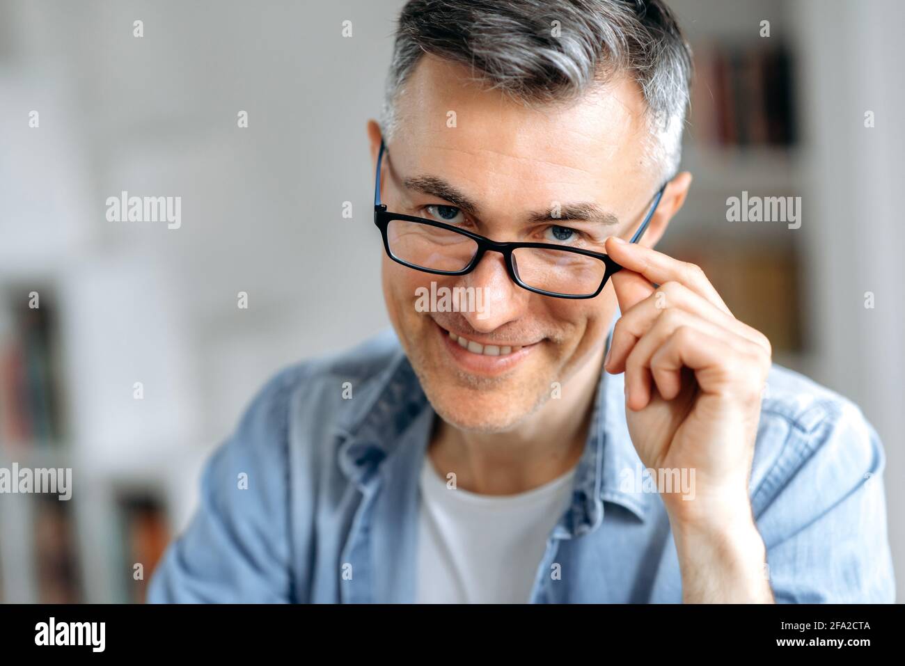 Primo piano ritratto riuscito sicuro influente uomo caucasico di mezza età con capelli grigi, in abiti casual eleganti, calare gli occhiali con la mano, su sfondo sfocato, guardando la macchina fotografica, sorridendo Foto Stock