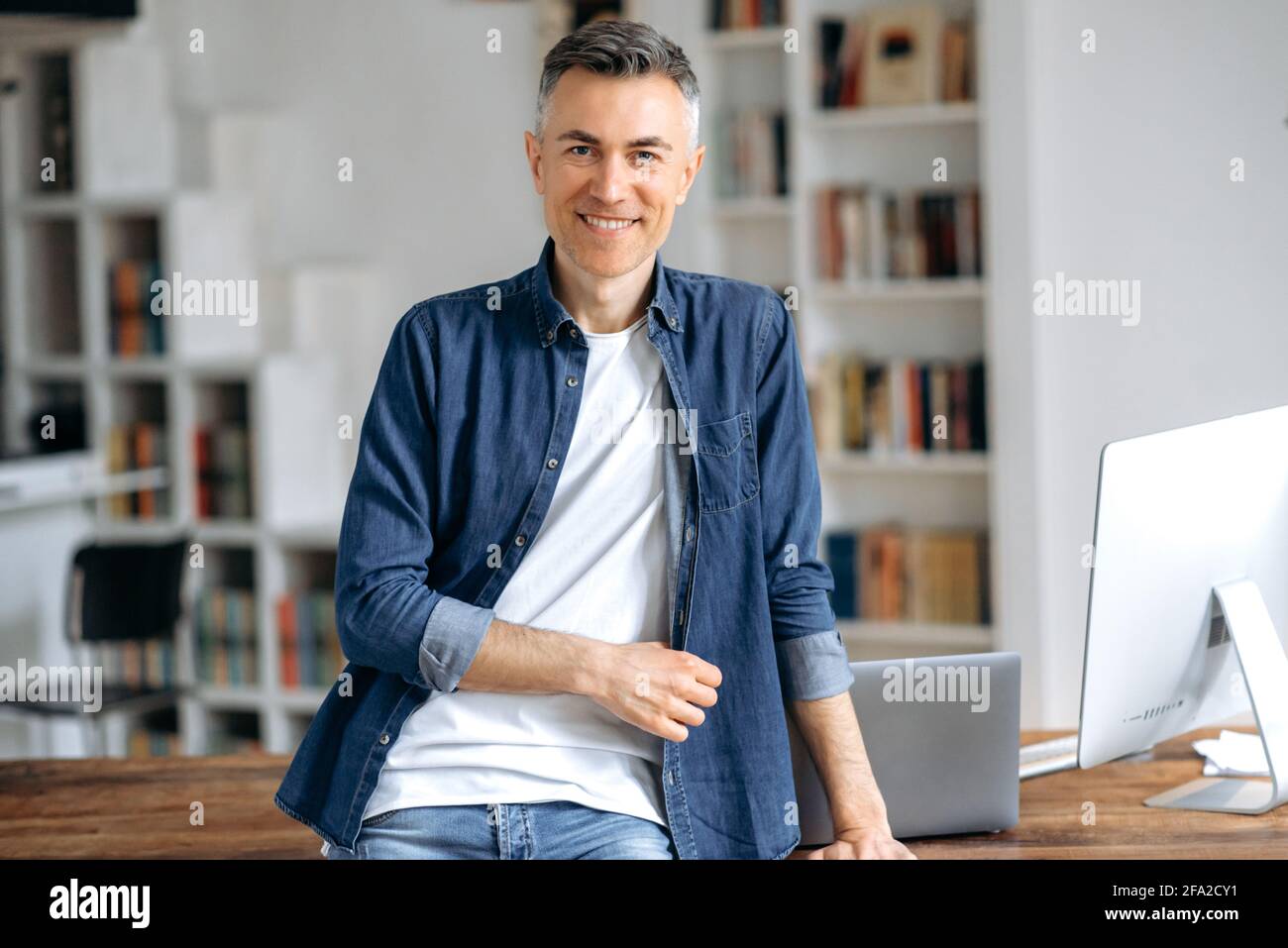 Ritratto di un bell'uomo caucasico con capelli grigi maturo di successo, in una camicia casual elegante in denim, si alza alla scrivania, guarda la fotocamera, sorride amichevole Foto Stock
