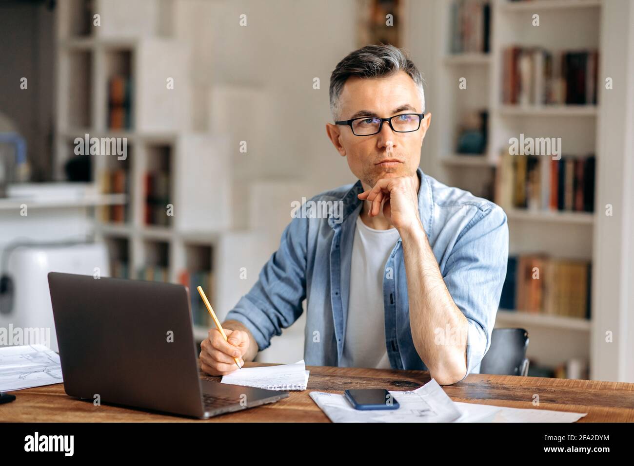Concentrato serio uomo d'affari caucasico o freelance con capelli grigi senior, indossando occhiali, lavorando con il laptop online, guardando via, pensando al business plan, sostenendo la testa con la mano Foto Stock