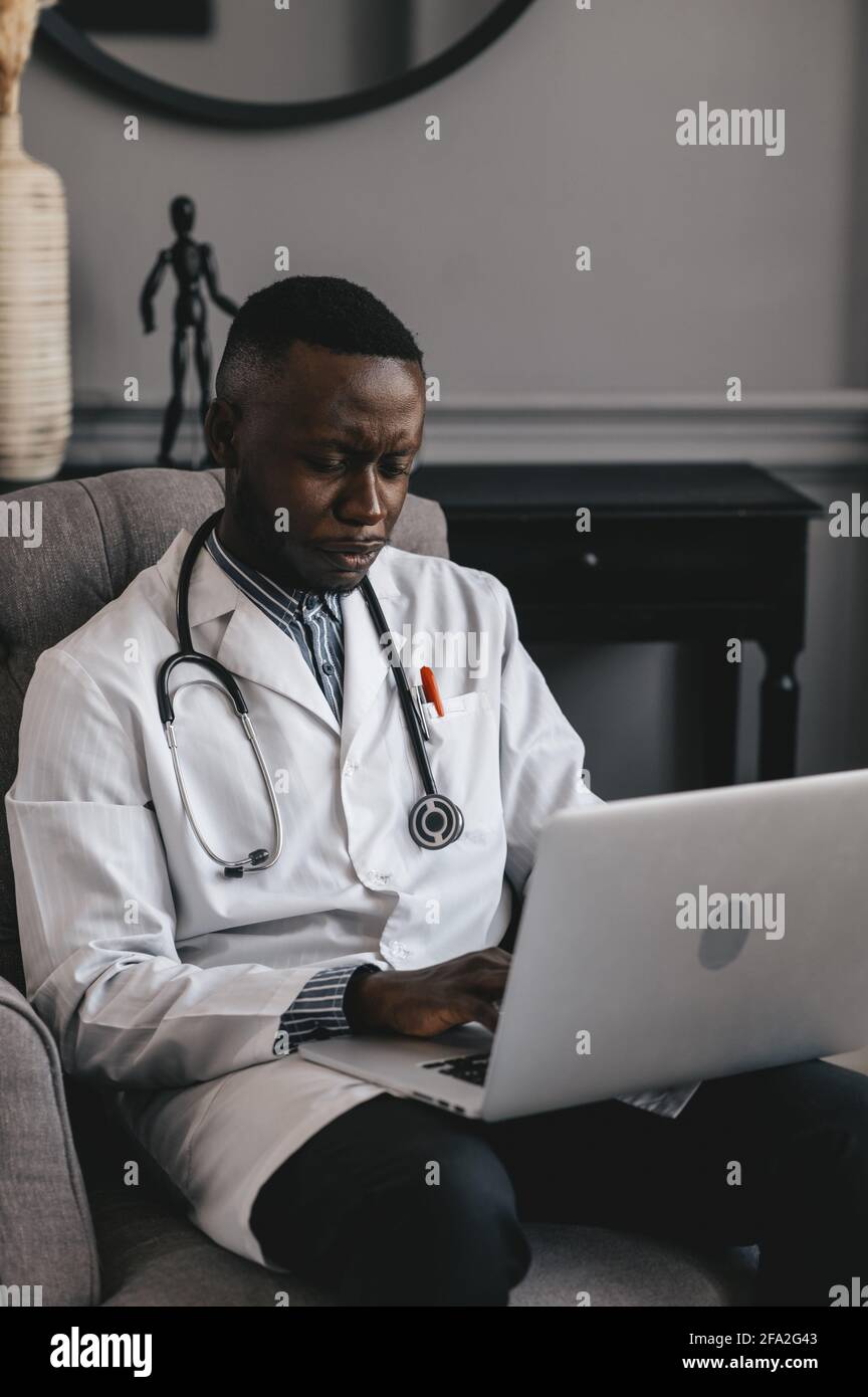 medico nero in una sedia grigia con un computer portatile a distanza consulta un paziente Foto Stock