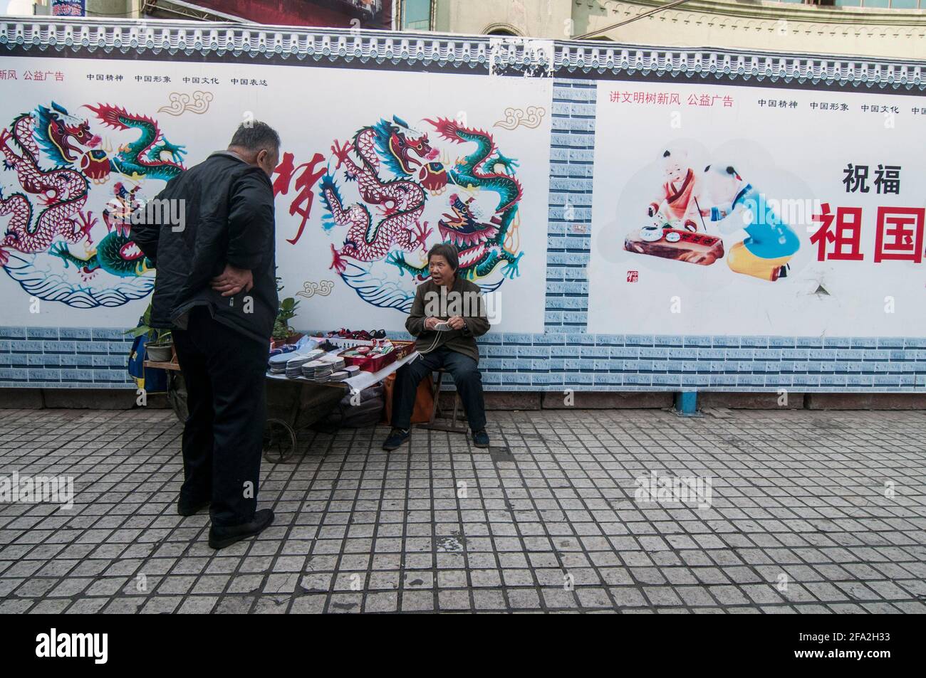 Una donna anziana chiacchiera con un uomo mentre siede infante di manifesti di propaganda su un muro dietro di lei, vendendo intarsi per scarpe e fissando il tessuto per le strade di Urumqi a Xinjiang, Cina, RPC. © Time-Snap Foto Stock