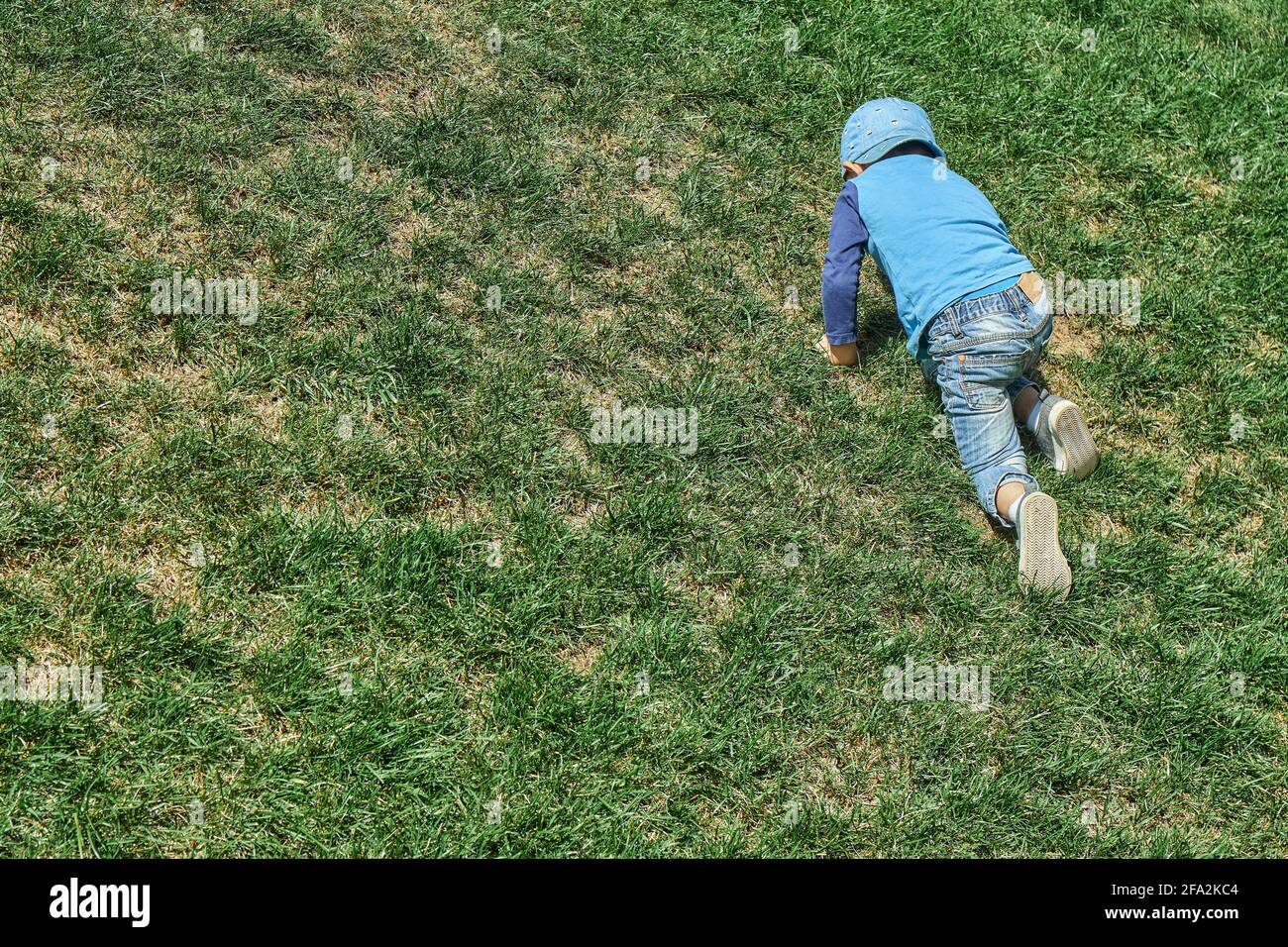 Un ragazzino giocoso in berretto blu sale su una ripida collina pendio coperto con erba verde lussureggiante in primavera parco su vista sul retro del giorno soleggiato Foto Stock